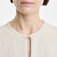 A woman is shown from the shoulders up against a white background, wearing the Maria McManus Pearl Button Blouse in light beige matte sateen Japanese Naia Renew, which features a distinctive pearl button at the collar, paired with silver earrings.