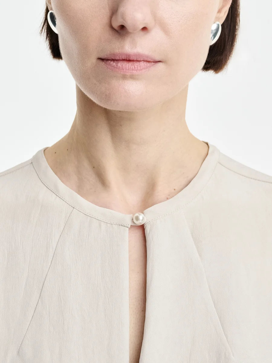 A woman is shown from the shoulders up against a white background, wearing the Maria McManus Pearl Button Blouse in light beige matte sateen Japanese Naia Renew, which features a distinctive pearl button at the collar, paired with silver earrings.