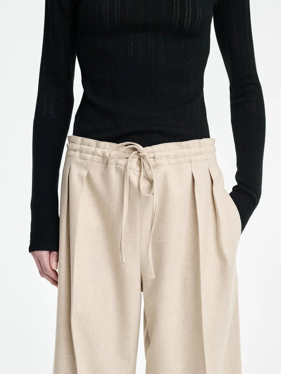 A person wears a black ribbed long-sleeve top paired with the Maria McManus Drawstring Trouser—a high-waisted beige wool-cashmere flannel pant with pleats and drawstring waist—styled for elevated loungewear; shown from shoulders to knees.