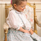 A young girl with braided hair sits on a floral-upholstered chair, holding the Antoinette Floral Blue Purse—its elegant pattern and hues complementing her gentle smile.