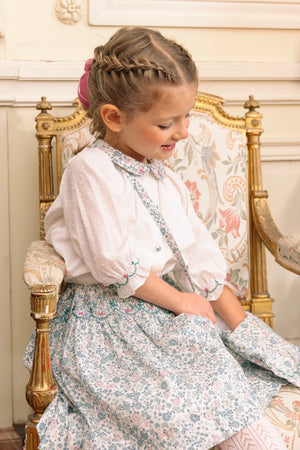 A young girl with braided hair sits on a floral-upholstered chair, holding the Antoinette Floral Blue Purse—its elegant pattern and hues complementing her gentle smile.