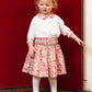 A young child with curly blond hair models the Antoinette Girls' Marie Red Floral Smocked Skirt and Blouse Set, featuring an embroidered collar, against a red upholstered background—a charming girls' holiday outfit.