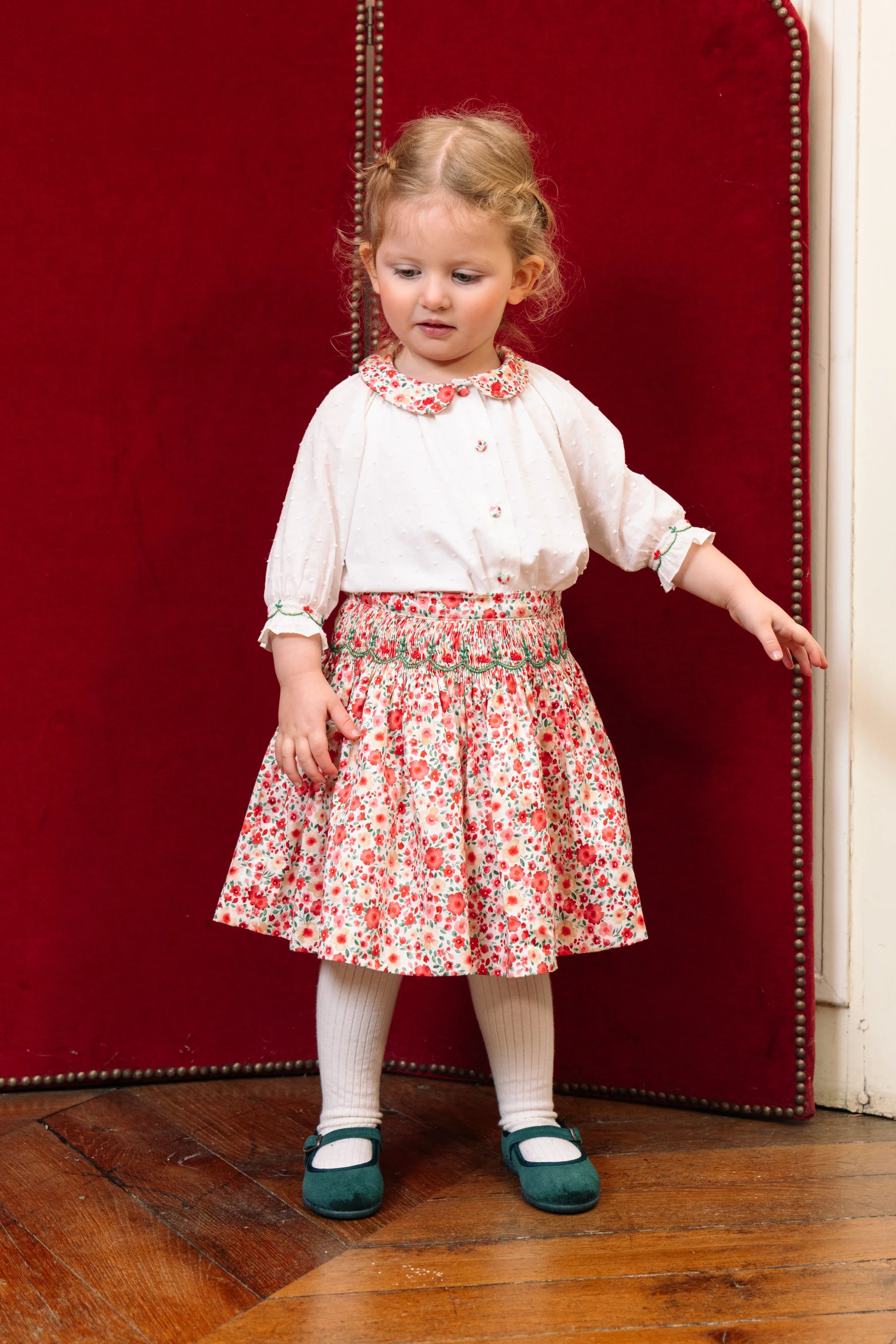 A young child with curly blond hair models the Antoinette Girls' Marie Red Floral Smocked Skirt and Blouse Set, featuring an embroidered collar, against a red upholstered background—a charming girls' holiday outfit.