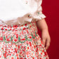 A girl wears the Antoinette Girls' Marie Red Floral Smocked Skirt and Blouse Set, standing against a solid red background for a charming holiday look.