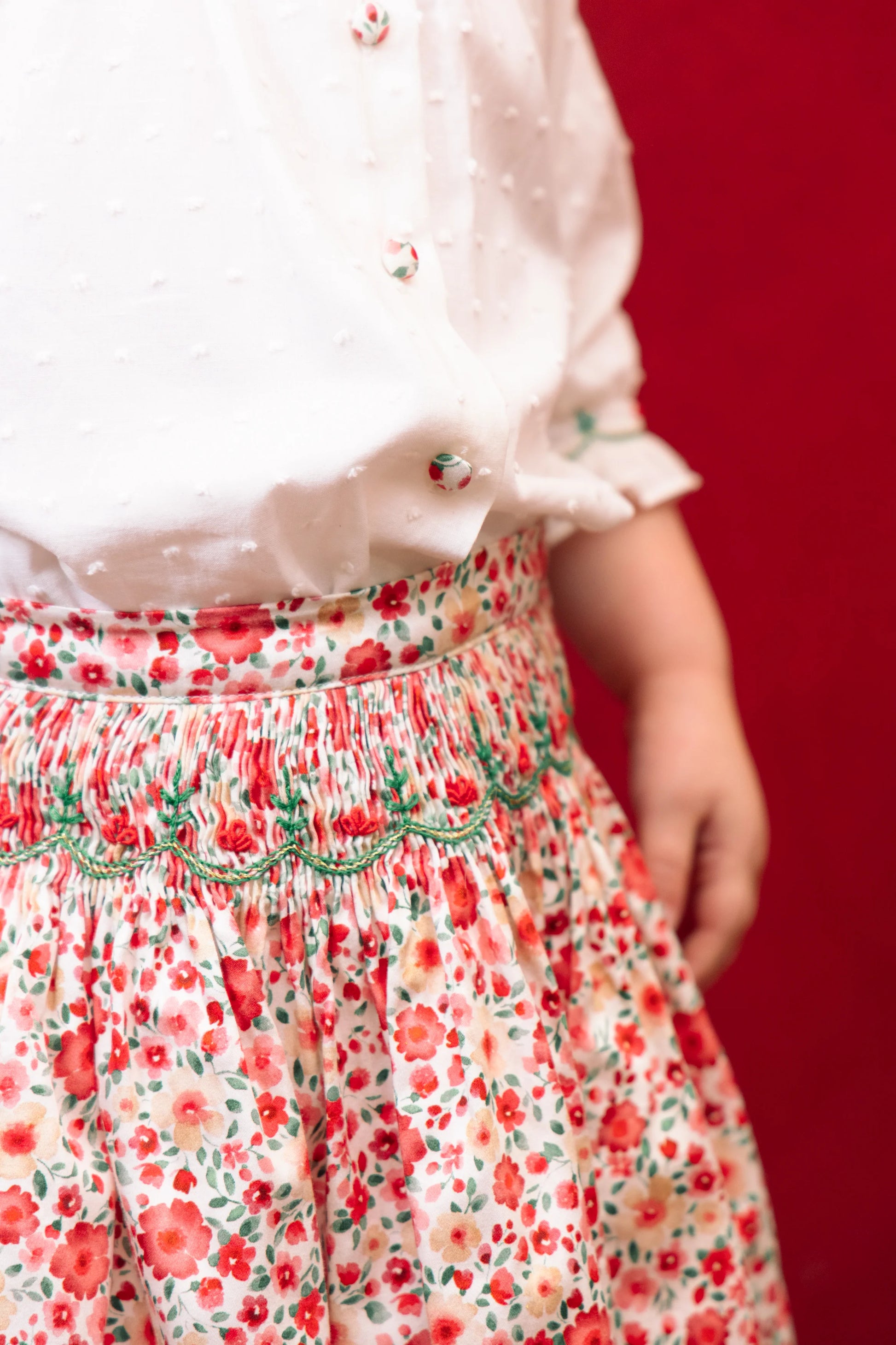 A girl wears the Antoinette Girls' Marie Red Floral Smocked Skirt and Blouse Set, standing against a solid red background for a charming holiday look.