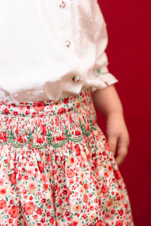 A girl wears the Antoinette Girls' Marie Red Floral Smocked Skirt and Blouse Set, standing against a solid red background for a charming holiday look.