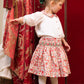 A young child wears the Antoinette Girls' Marie Red Floral Smocked Skirt and Blouse Set, standing on a wooden floor near an ornate red curtain, looking to the side with one arm extended.