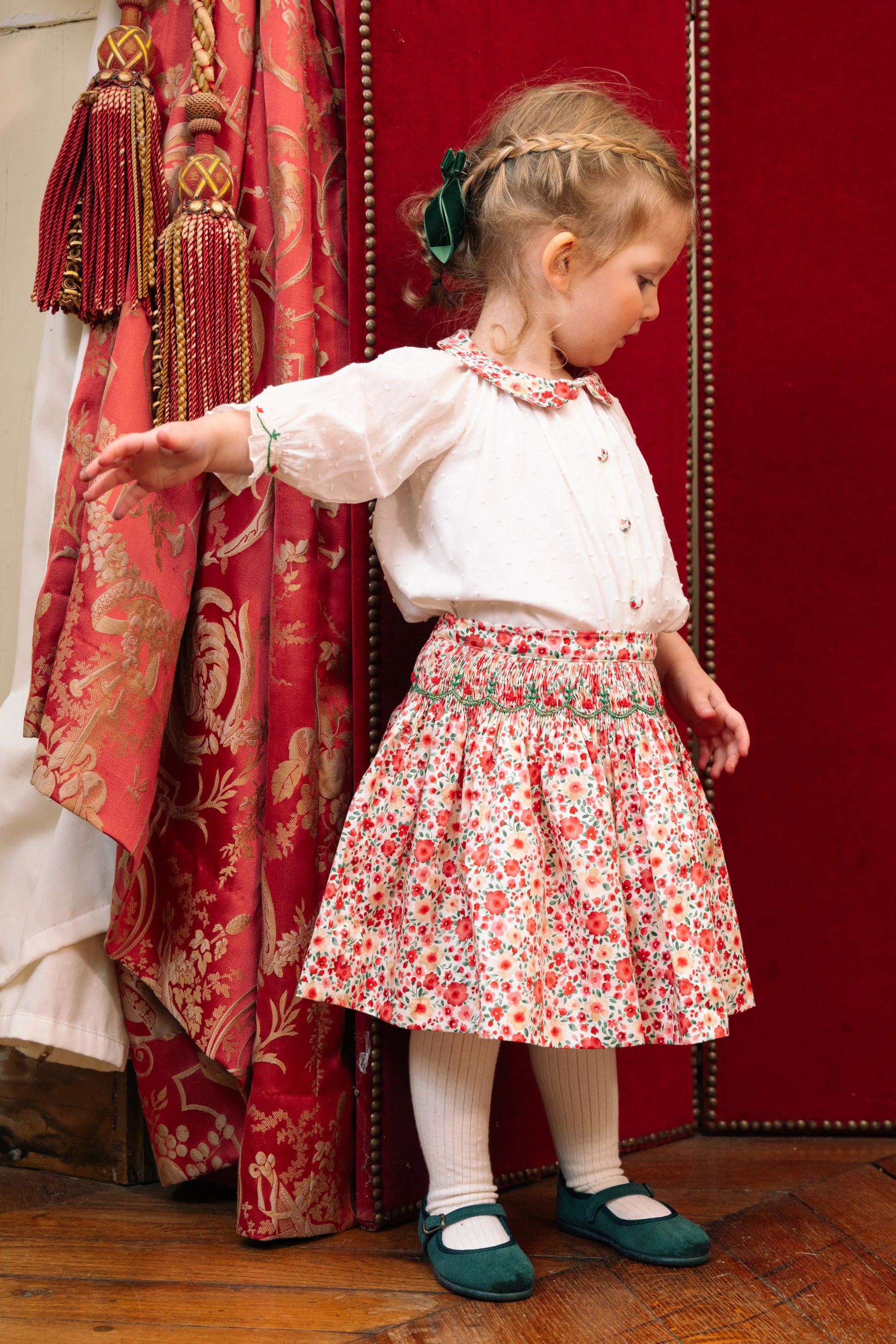 A young child wears the Antoinette Girls' Marie Red Floral Smocked Skirt and Blouse Set, standing on a wooden floor near an ornate red curtain, looking to the side with one arm extended.