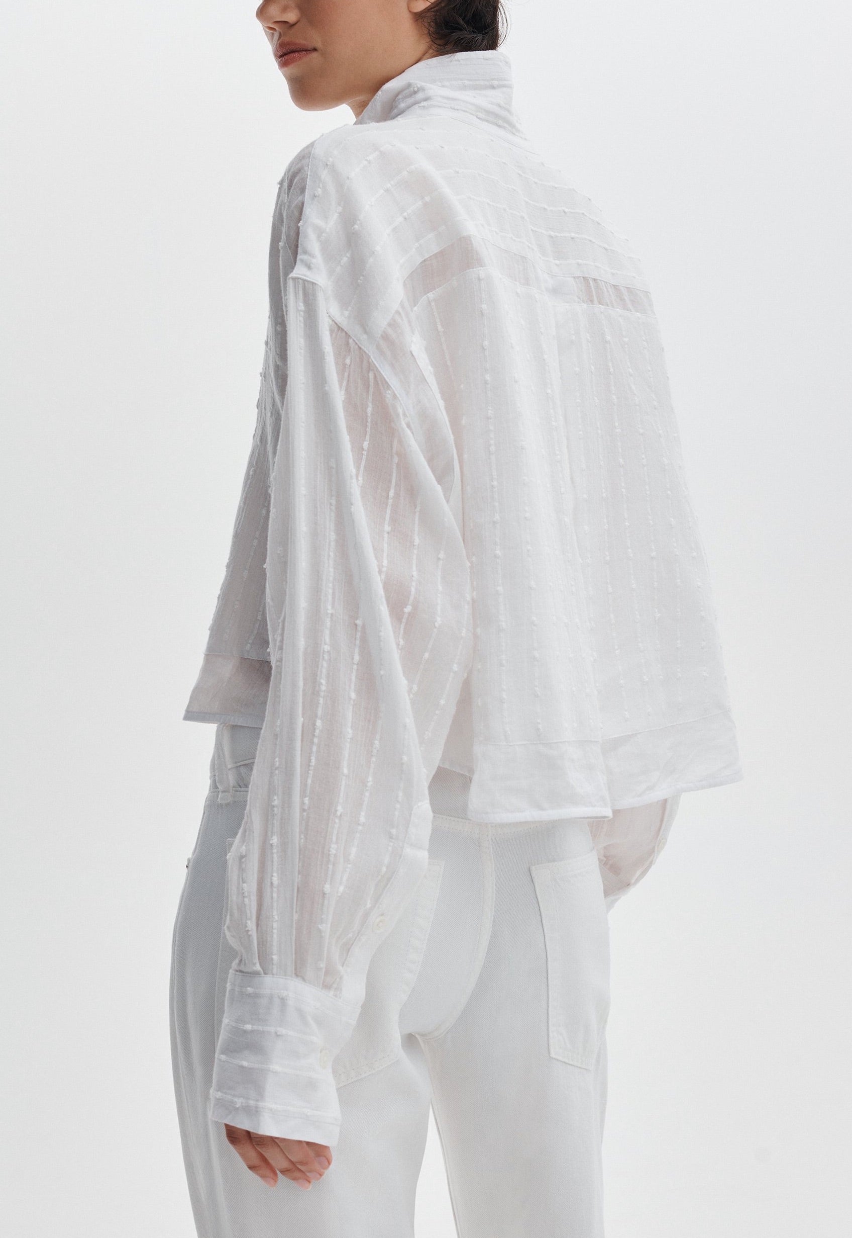 Model shown from the back wearing the Rag & Bone Marin Combo Shirt, a sheer white long-sleeve blouse with a textured pattern and relaxed fit, paired with white pants against a plain background.