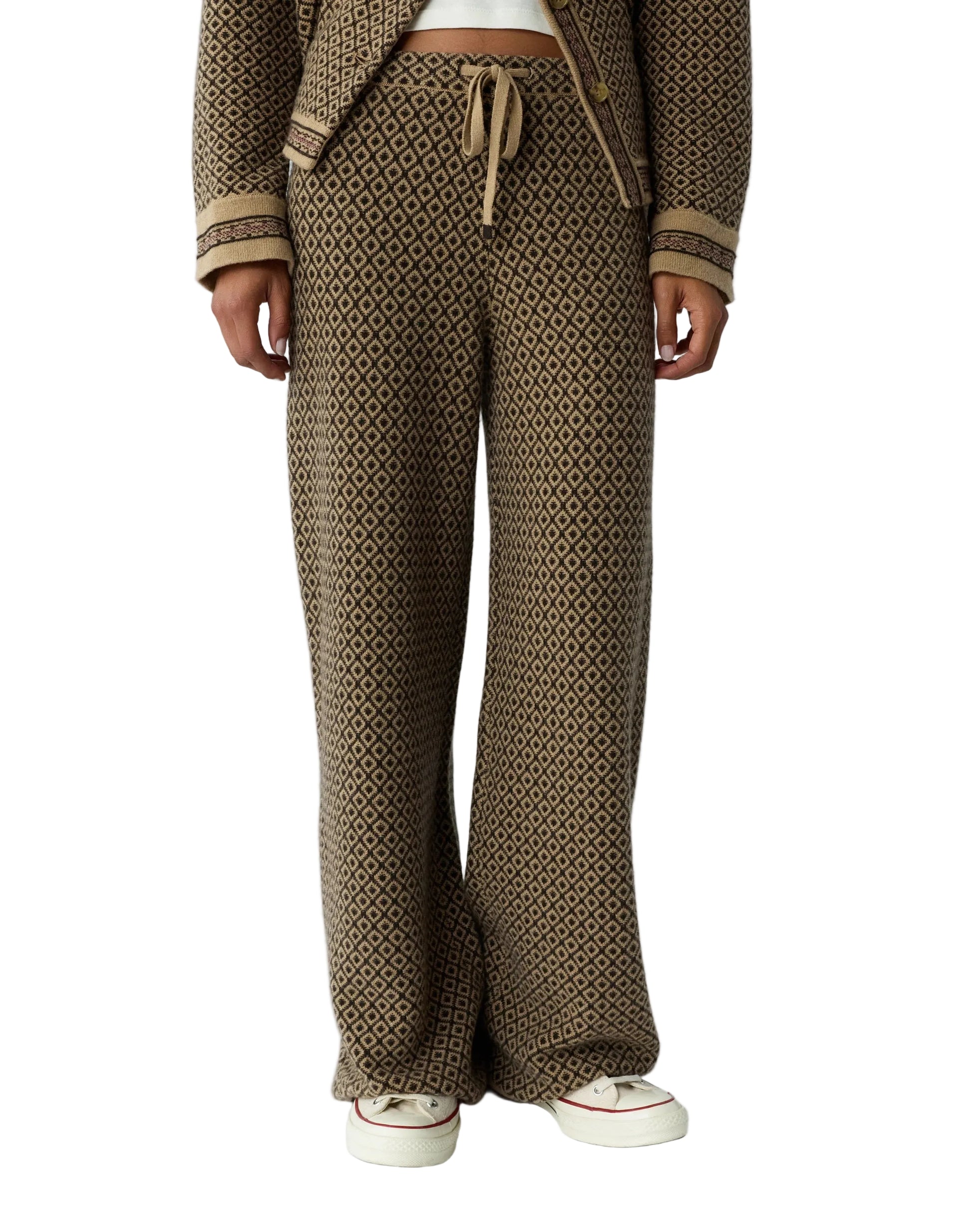 A person wearing the Marquise Pant by Guest in Residence—a brown patterned women’s cashmere pant with a straight leg and drawstring waist—paired with a matching jacket and white sneakers, stands against a white background.