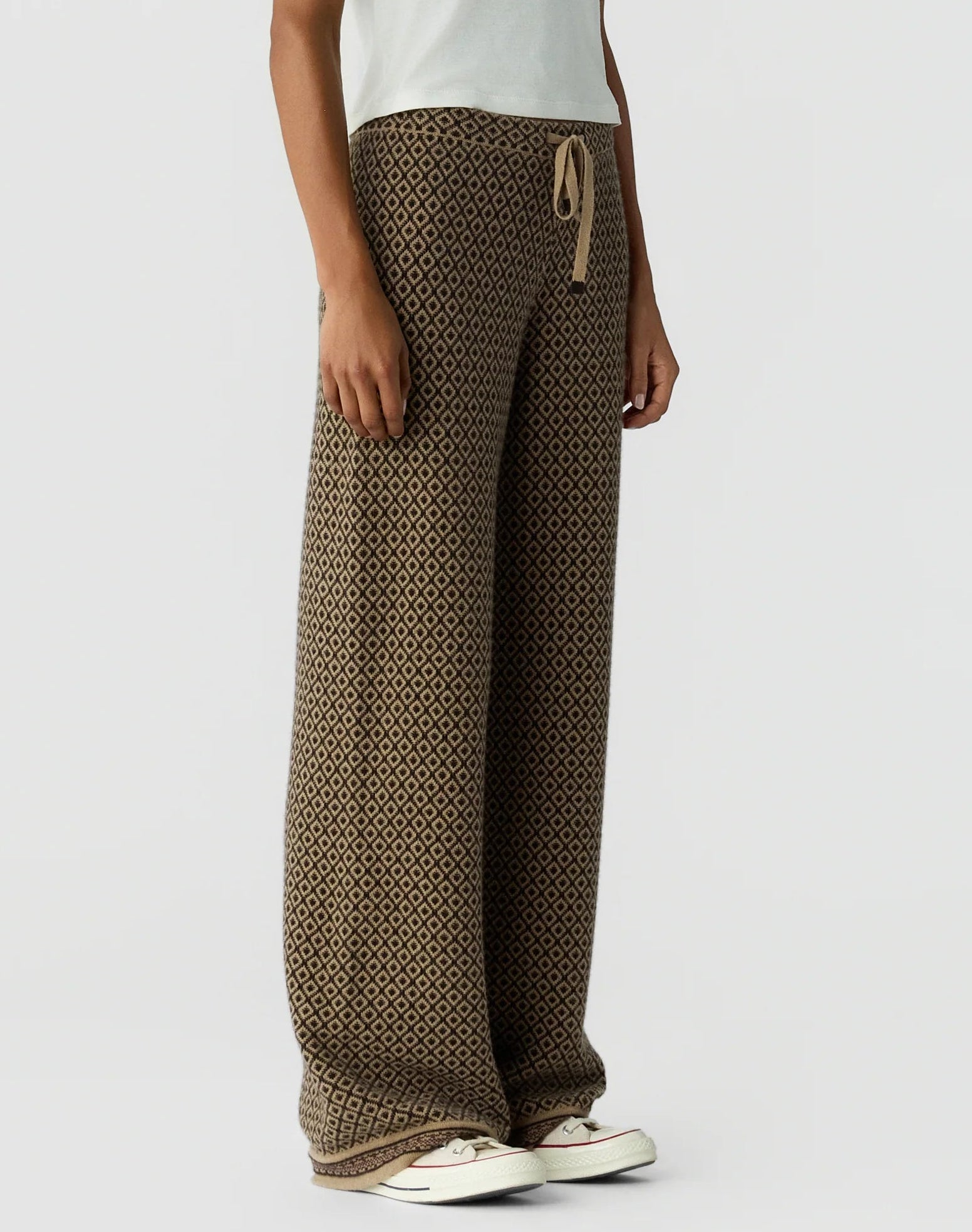 A person is wearing the Marquise Pant by Guest in Residence—loose brown patterned pants with a drawstring waist and straight leg—paired with a white T-shirt and white sneakers, standing against a plain light background.