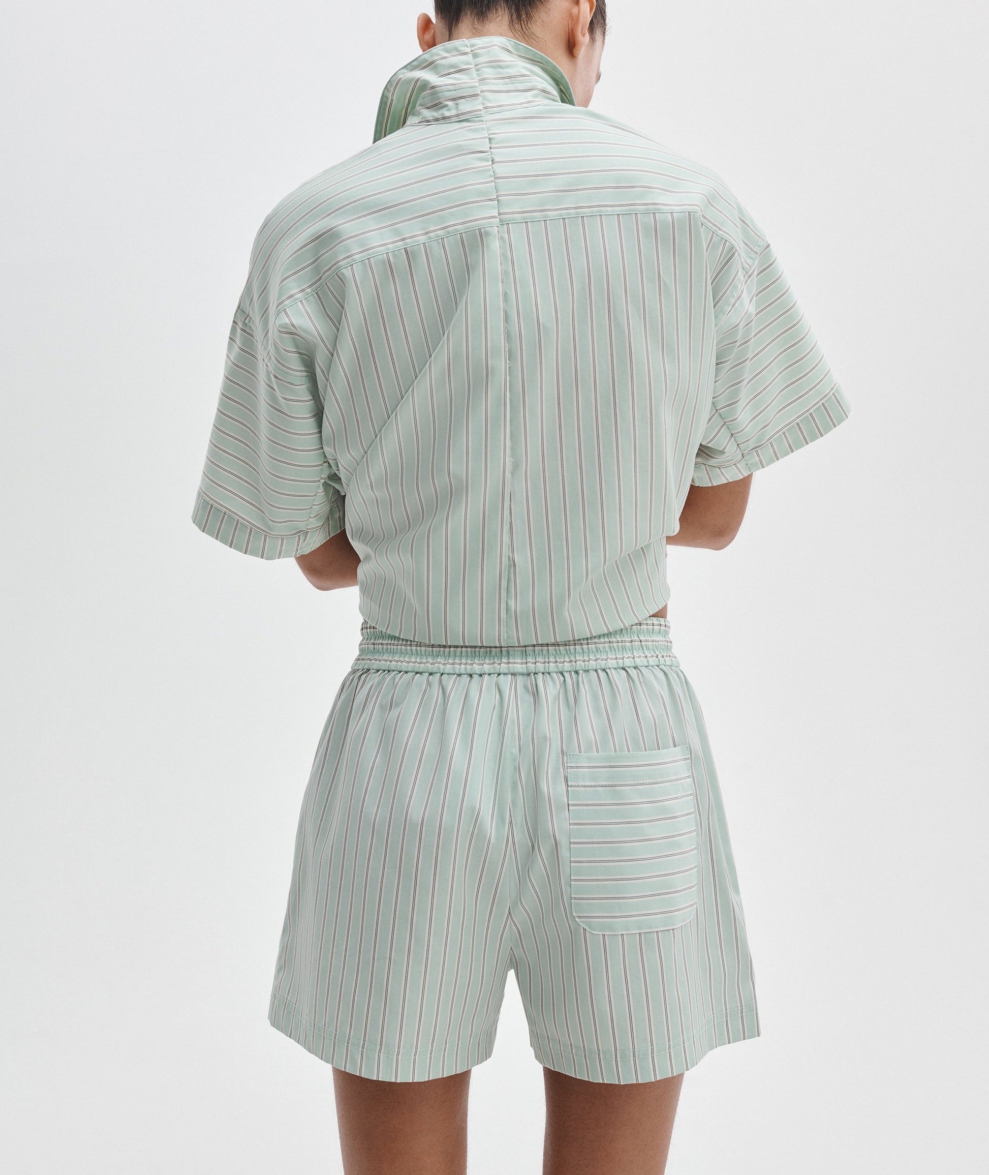Person facing away from the camera wearing the Rag & Bone Marvin Stripe Short, a light green, short-sleeved, collared romper with vertical stripes, adjustable waist, and elastic waistband.