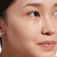 A woman with clear skin and a silver ear cuff rests her chin on her hand, gazing upward. A swatch of Supergoop Mineral Mattescreen SPF 40, a pore-blurring mattifying tinted sunscreen, is applied to her cheek.
