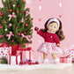 The limited edition Corolle Pearl Heart Petals Doll, dressed in a charming pink and red outfit, stands beside a beautifully adorned Christmas tree. Her silky brown hair perfectly complements the pink and red ornaments, along with the heart-patterned presents nestled under the tree.