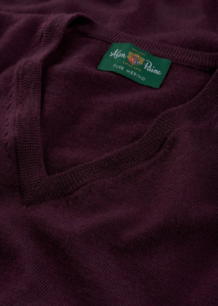 Close-up of a folded dark purple Millbreck V-Neck Merino Wool Sweater with a green label that reads “Alan Paine, Pure Merino.” This classic men’s layering piece by Alan Paine is expertly crafted from premium merino knitwear, offering unparalleled comfort and timeless style.