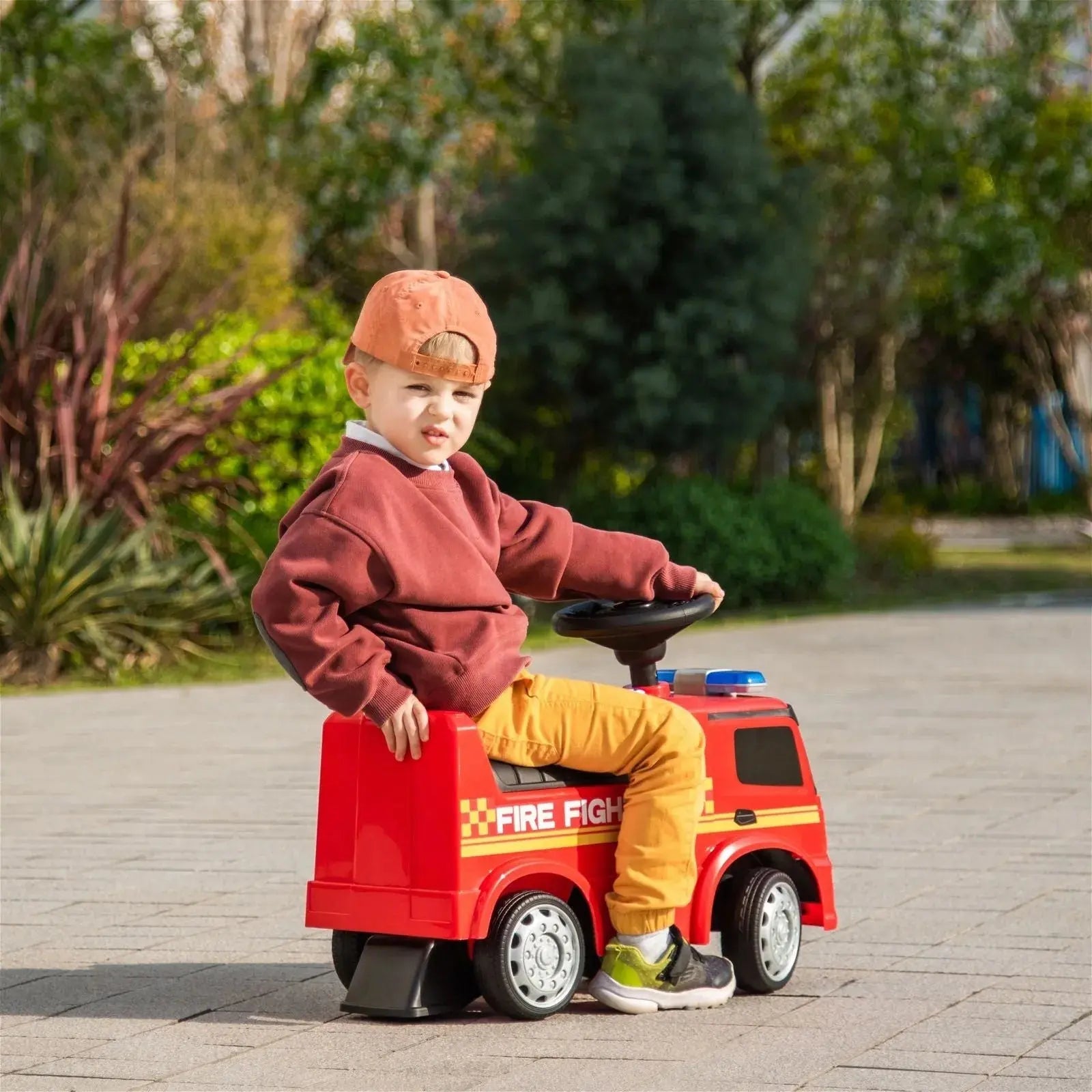 A young boy wearing a cap sits on a vibrant red Freddo Mercedes Antos Foot-To-Floor Fire Truck Ride-On in an outdoor area with paved ground and greenery in the background.