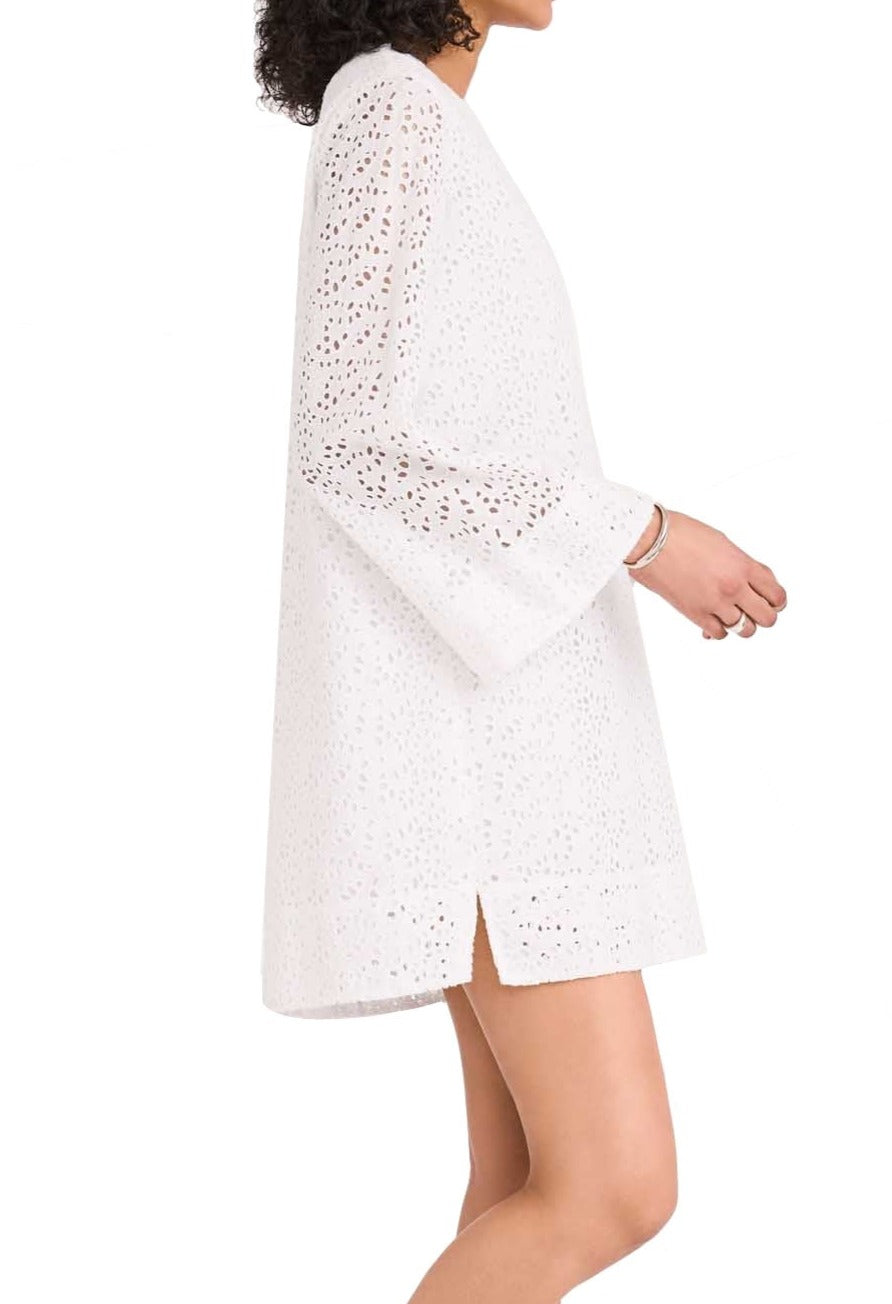 Woman wearing a white Merlette Charme dress from Merlette New York with a perforated pattern and bell sleeves, posing sideways.