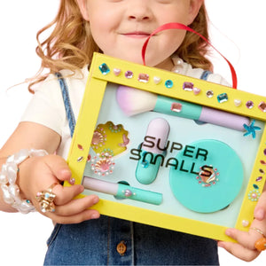 A child holds a yellow "Super Smalls" box containing the Mermaid Makeup Play Kit, a kids makeup set decorated with colorful gems—perfect for imaginative mermaid beauty adventures.