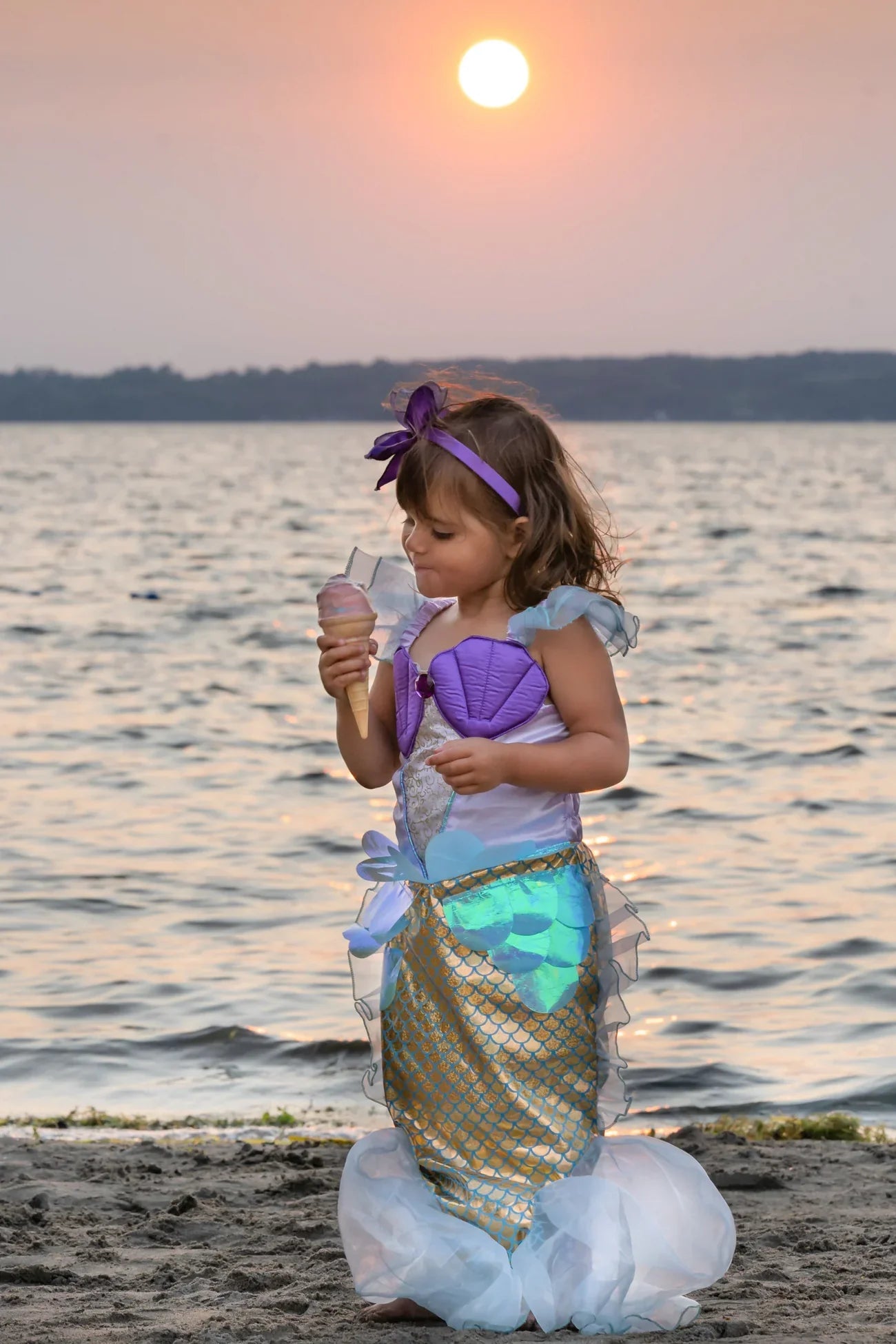 A child in a Great Pretenders Mermaid Dress & Headband delights in ice cream on the beach at sunset.