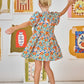 A young girl with blonde hair in a bun wears the Hunter Bell NYC Girls' Merritt Dress featuring a ruffled neckline, paired with yellow socks and mismatched sandals, standing with arms outstretched before framed wall art.