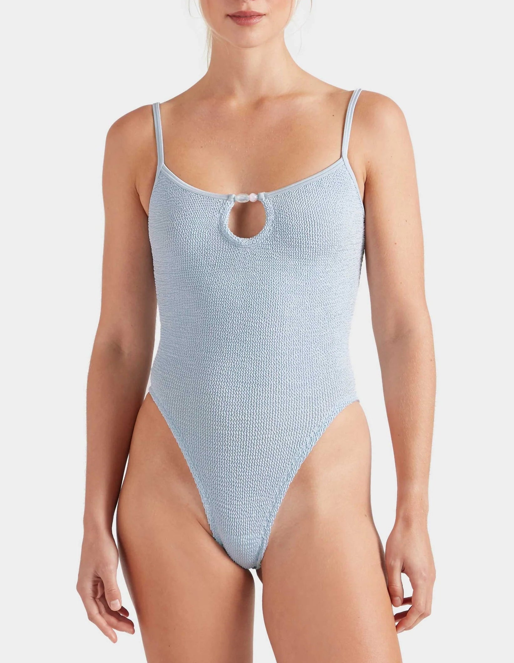 A woman wearing the Hunza G Mia Beaded Swim, a light blue crinkle fabric swimsuit with thin straps, a small front keyhole cutout, and a supportive fit designed to adapt to one size.