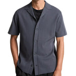 A person wears the vince Milano Shortsleeve Cabana in dark gray over a black t-shirt, standing with hands in pockets against a white background.