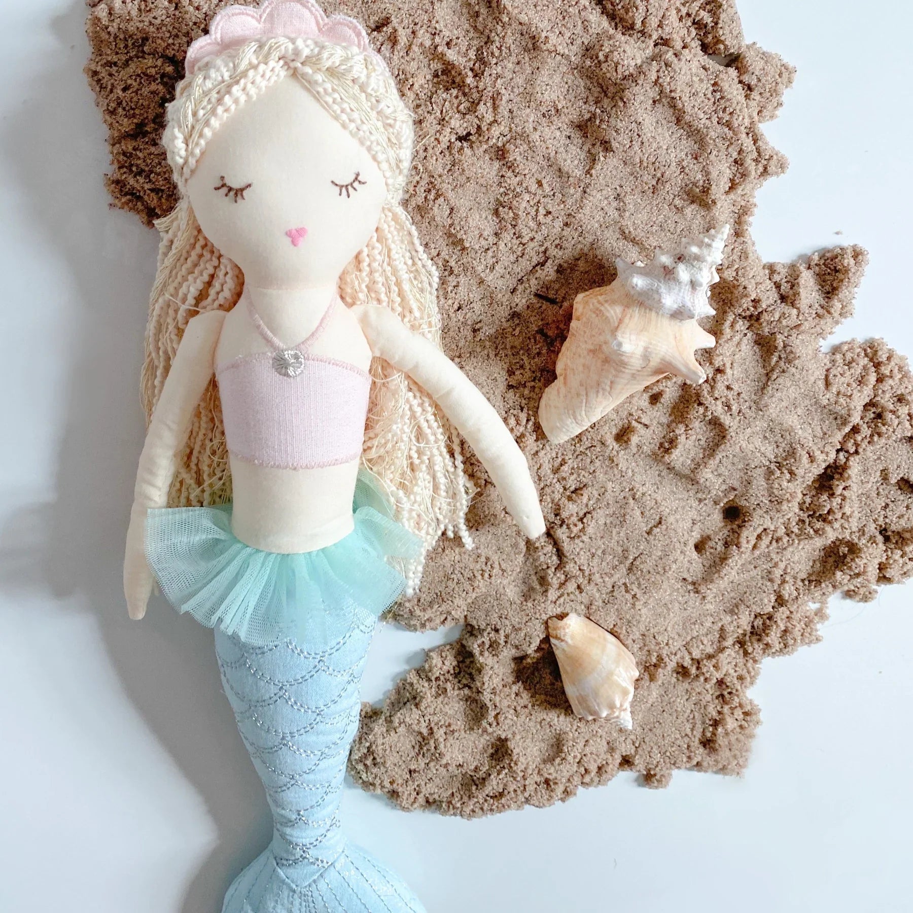 The MON AMI Mimi Mermaid Large by Mon Ami features a delightful mermaid doll with a light pink top and blue tail, resting on sandy textured material alongside two seashells. This enchanting figure is perfect for pretend play, adding a touch of ocean wonder to any adventure.