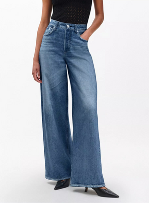 A person stands against a plain background wearing Rag & Bone’s Miramar Sofie Ankle denim look pants, a sleeveless black top, and black pointed-toe shoes.