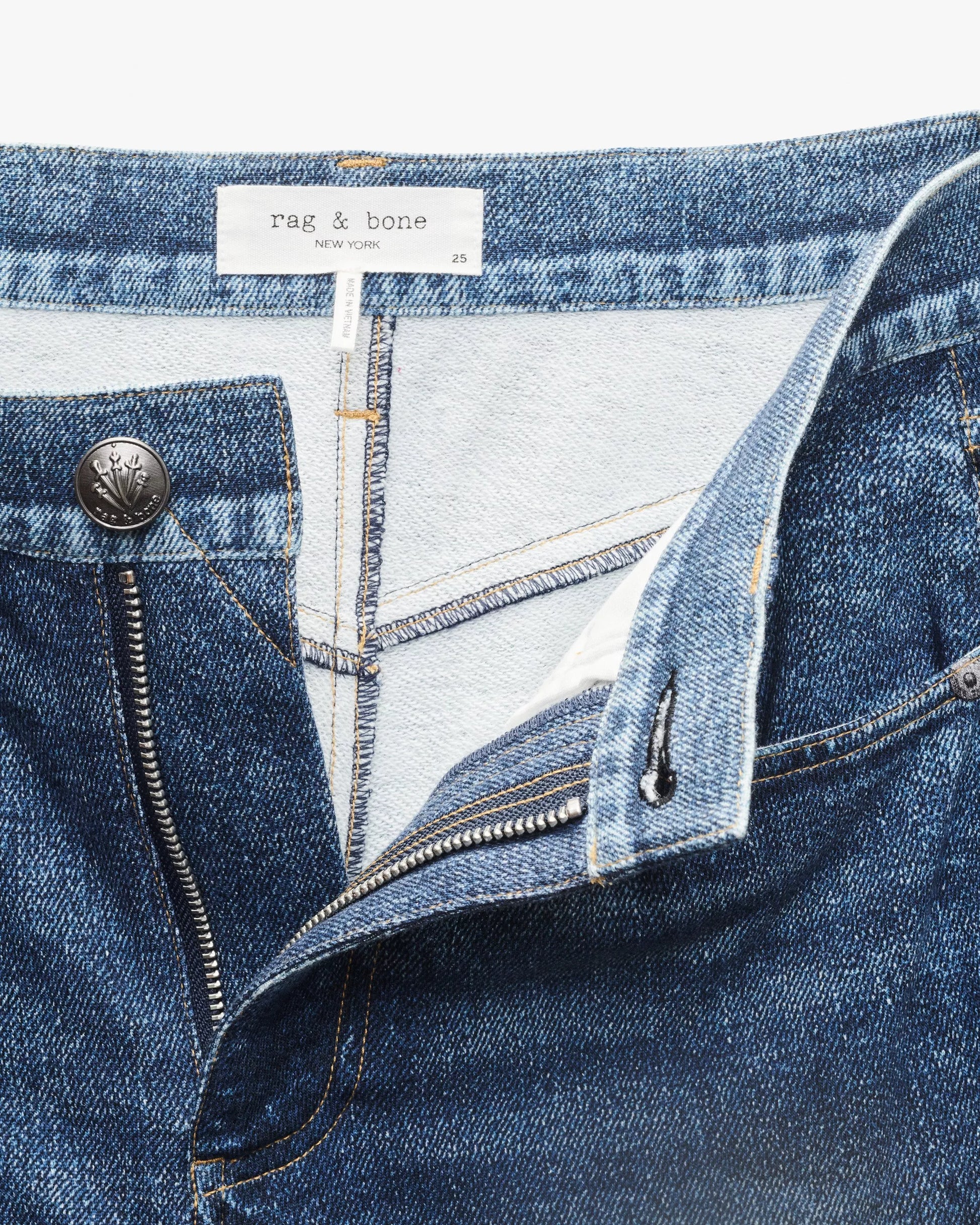 Close-up of blue denim-look Miramar Sofie Ankle pants by Rag & Bone, featuring an open zipper, waistband, label, and metal button.