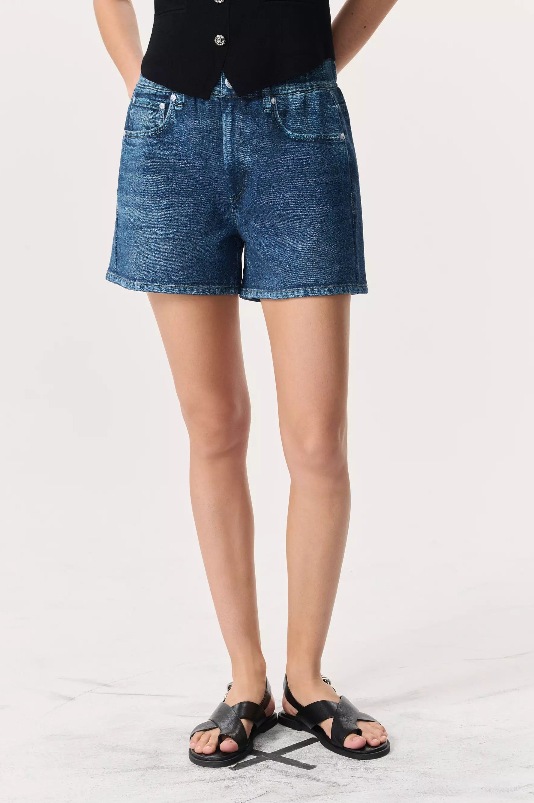 A person wearing Rag & Bone Miramar Terry Walking Shorts with a pull-on elastic waistband, paired with a black sleeveless top and black sandals, stands against a plain light background.