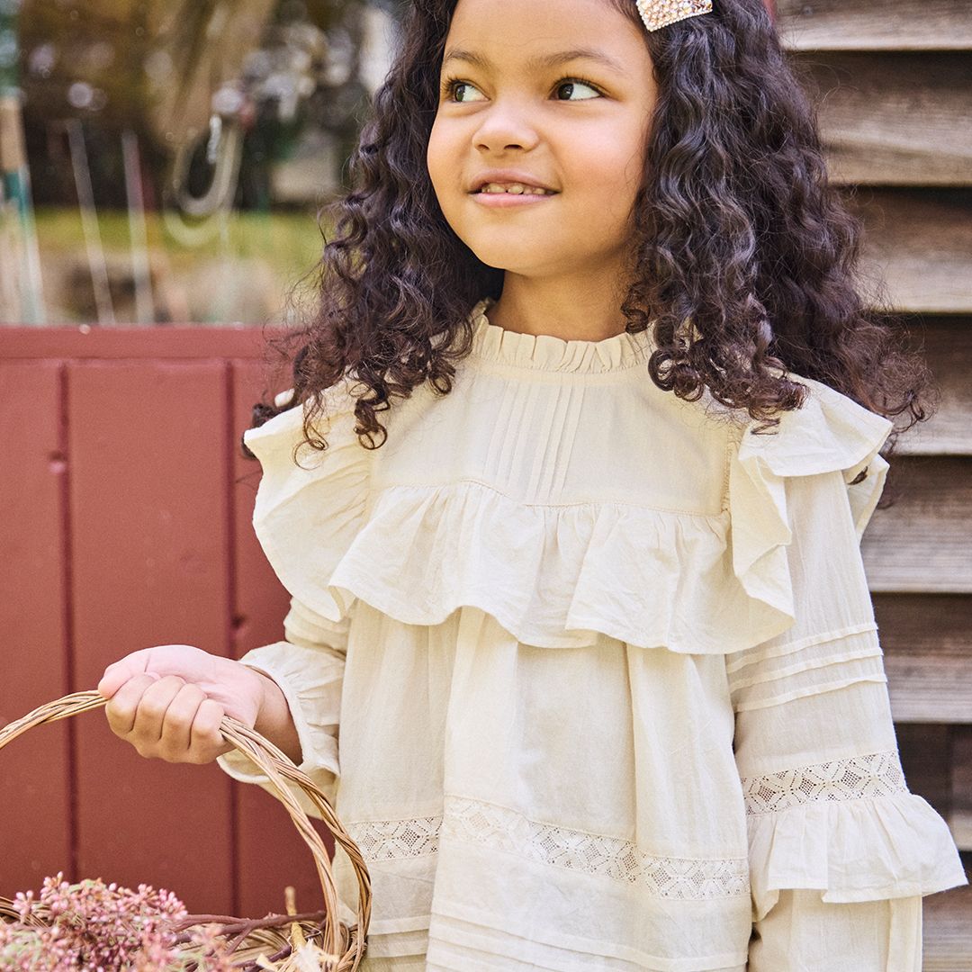 A girl in a Pink Chicken Girls' Lace Mira Top, holding a basket, looks incredibly charming in her 100% cotton cambric outfit.