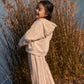 Wearing the Louise Misha Girls’ Osalia Sweatshirt—a beige fleece hoodie adorned with delicate embroidery—a young girl smiles by tall grass near a body of water on a sunny day.