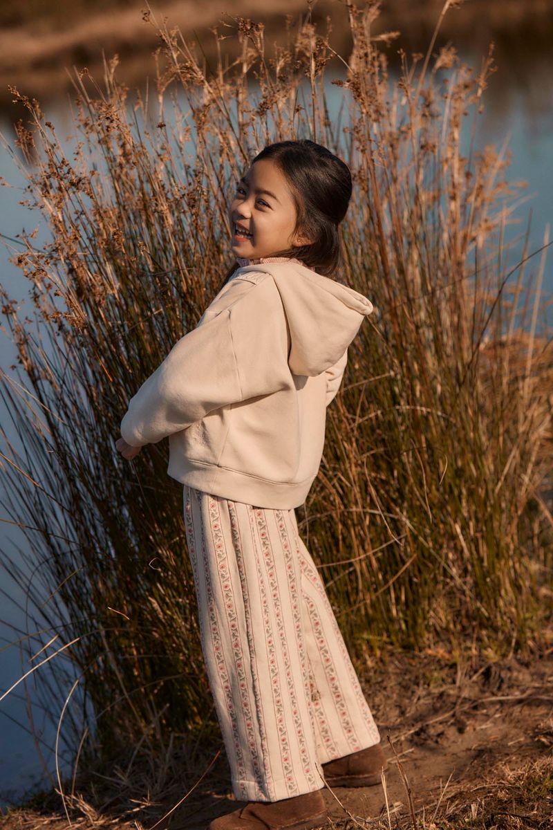 Wearing the Louise Misha Girls’ Osalia Sweatshirt—a beige fleece hoodie adorned with delicate embroidery—a young girl smiles by tall grass near a body of water on a sunny day.