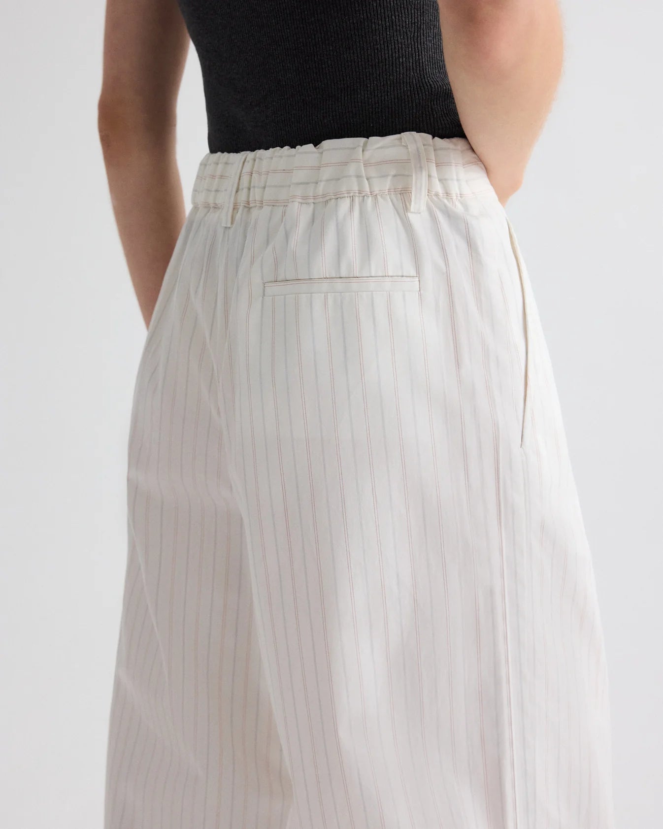 A person wears TWP’s Hudson St. Cotton Stripes Pant—a high-waisted, wide-leg white pinstripe cotton poplin trouser—paired with a sleeveless black top, shown from the waist down with hands behind their back.