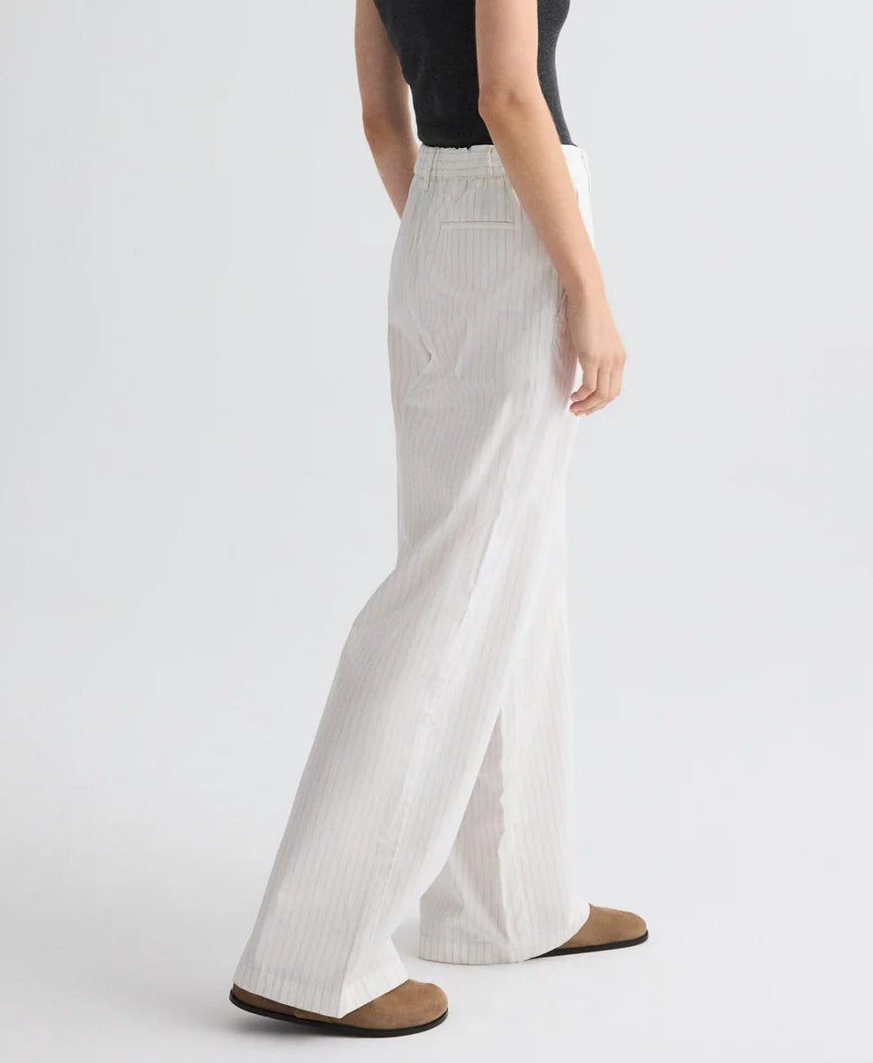 A person stands against a plain light background wearing TWP's Hudson St. Cotton Stripes Pant in white pinstriped cotton poplin, paired with a black sleeveless top and brown flat shoes.