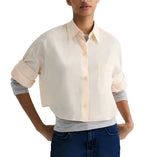 A person wearing the TWP Next Ex Superfine Cotton Shirt—a light-colored, short-sleeve button-down with a cropped boxy silhouette—over a gray long-sleeve top and blue jeans, standing with hands on their hips.