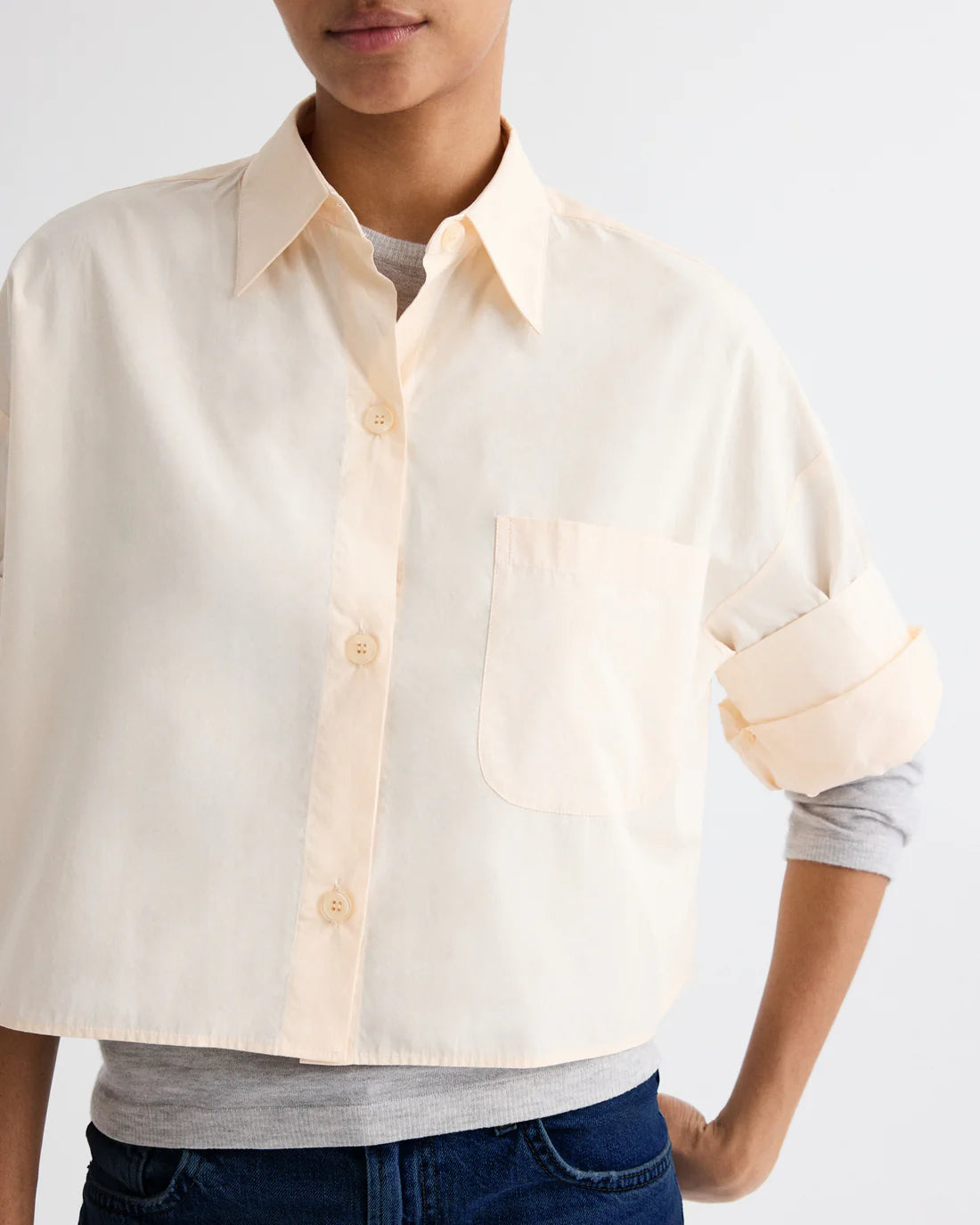 A person wears the TWP Next Ex Superfine Cotton Shirt—a light-colored, cropped boxy Italian cotton style with chest pocket—over a gray long-sleeve shirt and blue jeans, standing against a plain background.