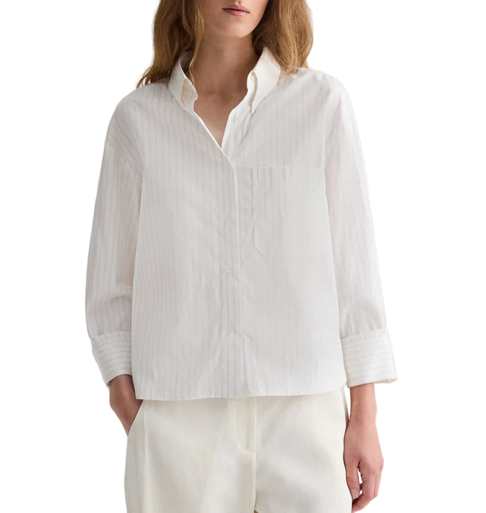 A person wears the TWP J.D. Striped Shirt, a loose-fitting, long-sleeved cotton poplin shirt with subtle vertical stripes and a collar, paired with matching white pants. The person's head is partially out of frame.