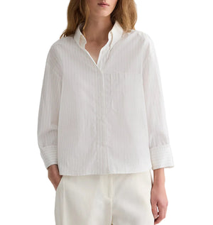A person wears the TWP J.D. Striped Shirt, a loose-fitting, long-sleeved cotton poplin shirt with subtle vertical stripes and a collar, paired with matching white pants. The person's head is partially out of frame.