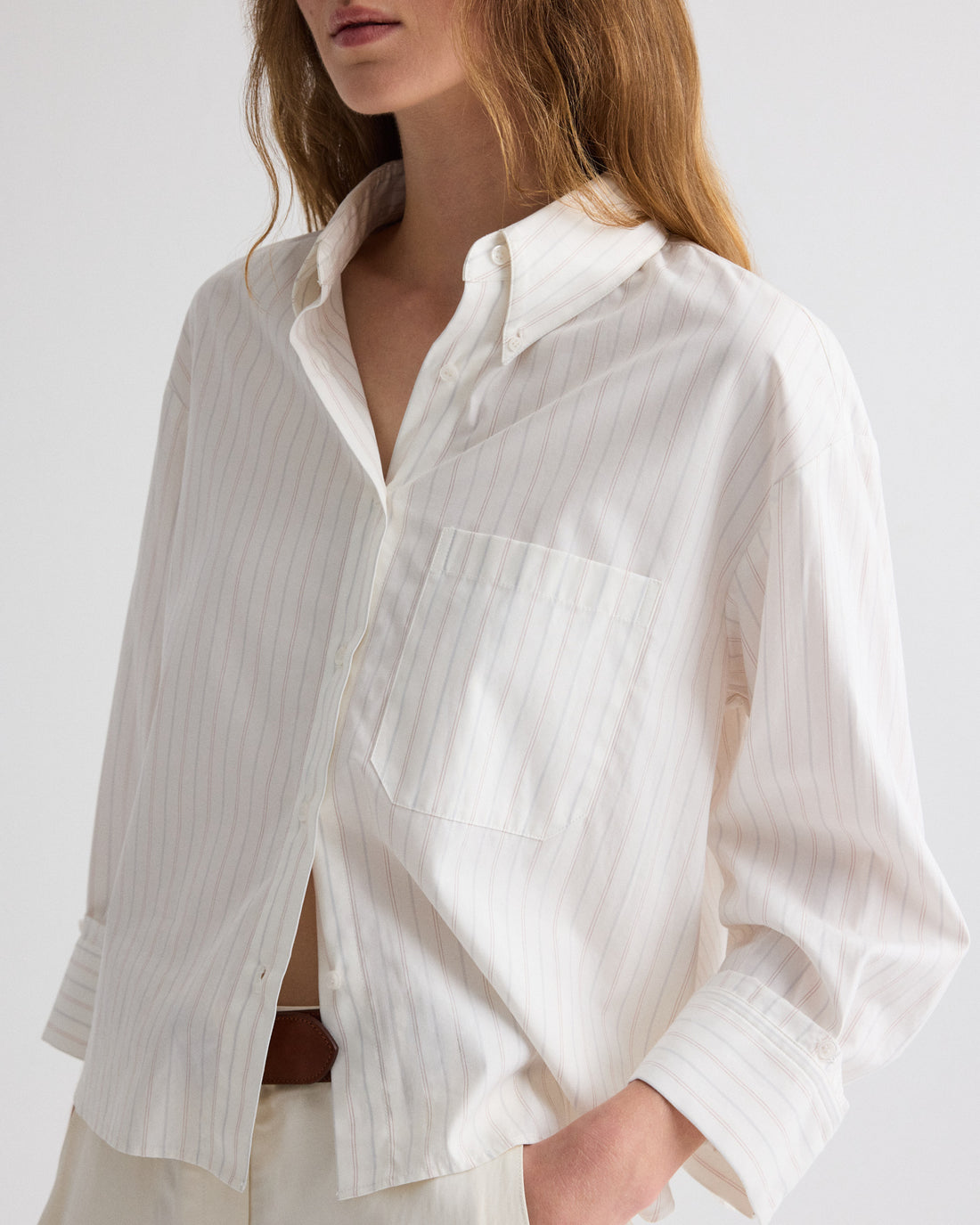 A person wearing TWP’s J.D. Striped Shirt—a loose-fitting, white button-down with thin vertical pinstripes—partially tucked into light-colored pants.