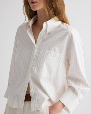 A person wearing TWP’s J.D. Striped Shirt—a loose-fitting, white button-down with thin vertical pinstripes—partially tucked into light-colored pants.