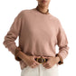 A person wears the TWP Boy Cashmere Crew in light pink over a brown collared shirt, paired with white pants and a brown belt with a gold buckle.