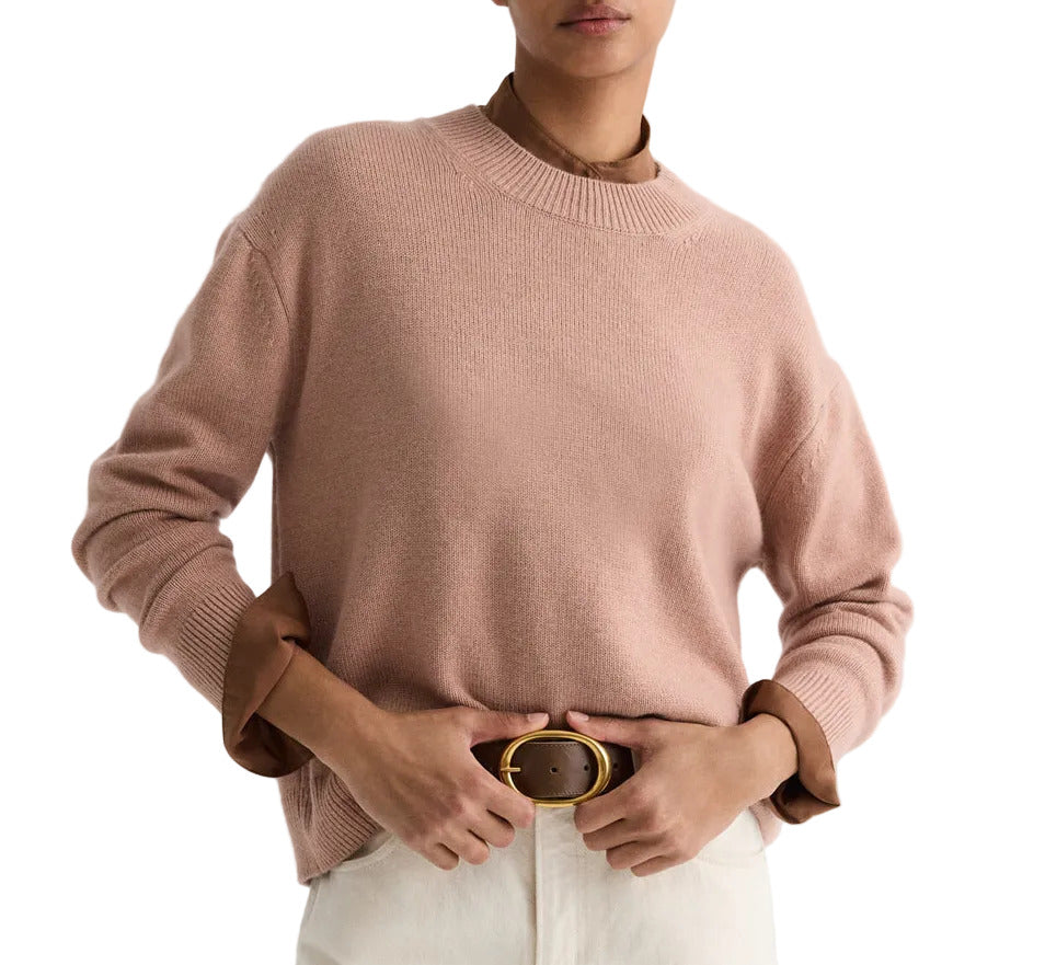 A person wears the TWP Boy Cashmere Crew in light pink over a brown collared shirt, paired with white pants and a brown belt with a gold buckle.