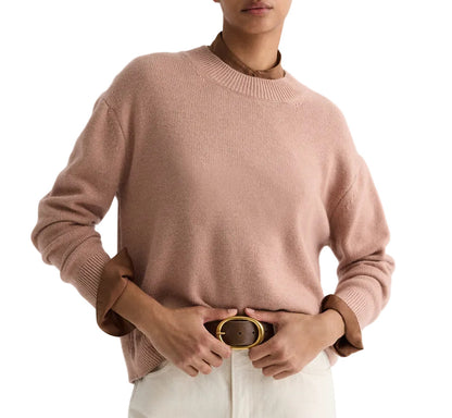 A person wears the TWP Boy Cashmere Crew in light pink over a brown collared shirt, paired with white pants and a brown belt with a gold buckle.