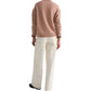 A person stands with their back to the camera, wearing the TWP Boy Cashmere Crew sweater in light brown, white wide-leg pants, and black shoes against a white background.