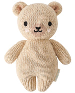 The Baby Honey Bear by Cuddle + Kind is an heirloom-quality beige teddy bear, hand-knit from Peruvian cotton yarn with black eyes, a small nose, and blushed cheeks, shown against a white background.