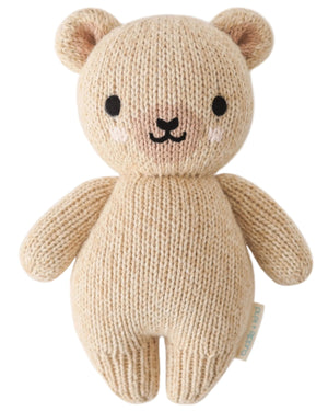 The Baby Honey Bear by Cuddle + Kind is an heirloom-quality beige teddy bear, hand-knit from Peruvian cotton yarn with black eyes, a small nose, and blushed cheeks, shown against a white background.
