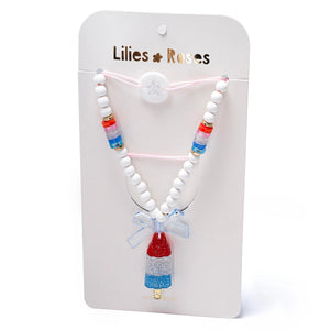 The Popsicle Rocket Red White Blue Necklace by Lillies & Roses features red, white, and blue beads with a rocket popsicle pendant, presented on a branded card.