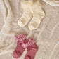 Two pairs of Noralee Ruffle Socks for children, featuring tulle ruffle trim in soft cotton blend—one cream and one pink—are displayed on a textured fabric background.