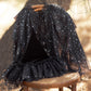 A Noralee Tulle Cape in black, adorned with silver stars, hangs on a chair outdoors—ideal for dress-up or special occasions. A coordinating black pointed hat and skirt are also displayed on the chair.